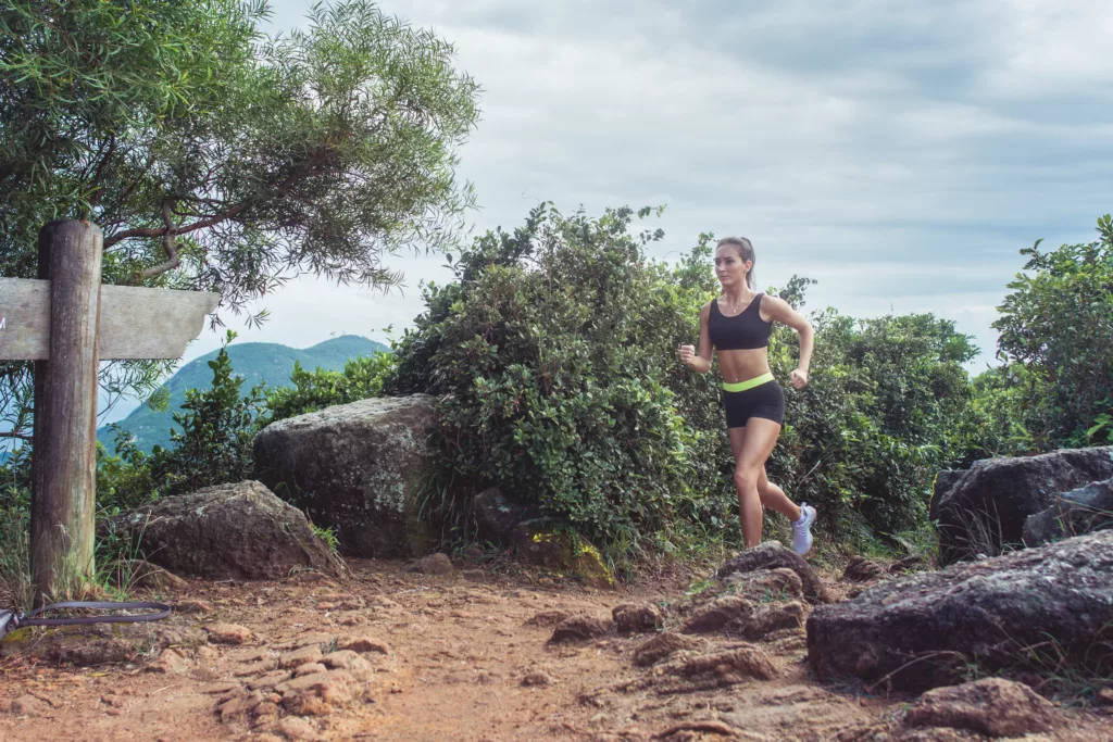 Young Sportswoman Cross Country Running On Dirty Rocky Footpath In Mountains In Summer. Fit Girl Jogging Outdoors In Countryside.